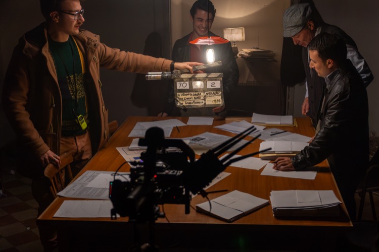 Il ciak davanti a un tavolo coperto di documenti, illuminato da una lampada, con gli attori in costume